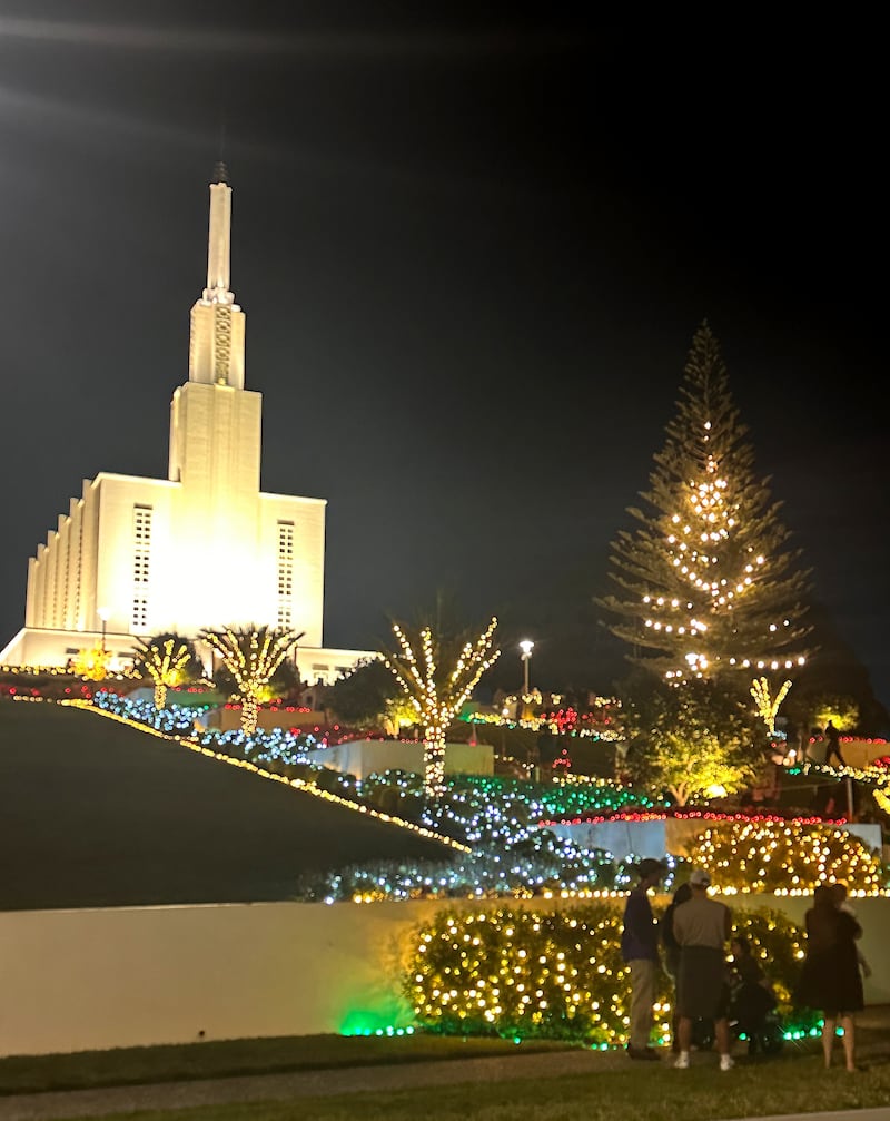 Los jardines del Templo de Hamilton, Nueva Zelanda, están iluminados con luces navideñas en esta foto de diciembre de 2025. En primer plano, los visitantes observan una escena de la Natividad frente al templo.