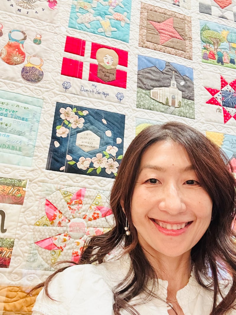 Sachiko Aldous está de pie junto a su bloque de colcha, que es azul con flores de cerezo aplicadas en la parte superior, en la exhibición "The Family Quilt" (La colcha familiar) en el Centro de Visitantes de Temple Square en Salt Lake City, julio de 2026.