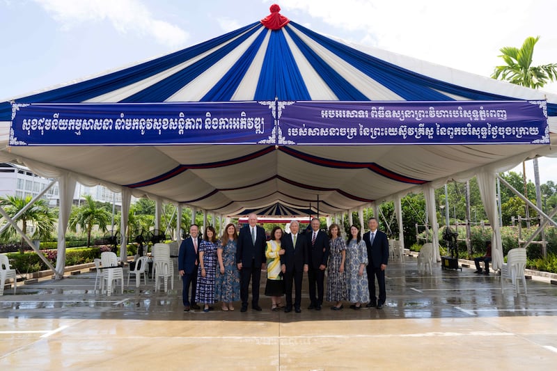 El élder Gerrit W. Gong y su esposa, la hermana Susan Gong, en el centro, se unen a cuatro Setentas Autoridades Generales y sus esposas bajo una carpa proporcionada por el gobierno de Camboya para el primer día de los recorridos de casa abierta del Templo de Phnom Penh Camboya el miércoles 12 de agosto de 2026. El letrero dice: "La unidad en la religión trae paz y desarrollo. Felicidades por el Templo de Phnom Penh Camboya de La Iglesia de Jesucristo de los Santos de los Últimos Días".
