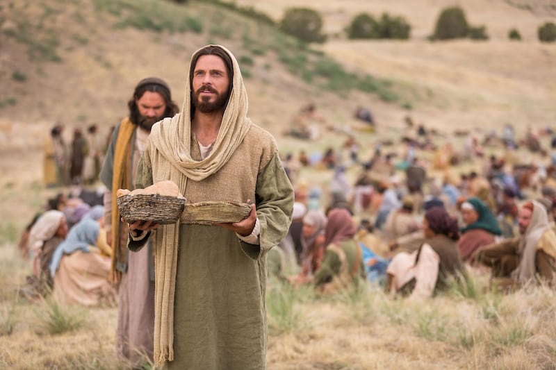 Jesucristo sosteniendo canastas con pan y peces, con muchas personas detrás de Él.