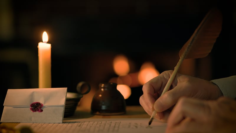 Un hombre escribe una carta con una pluma, con un fuego encendido al fondo.