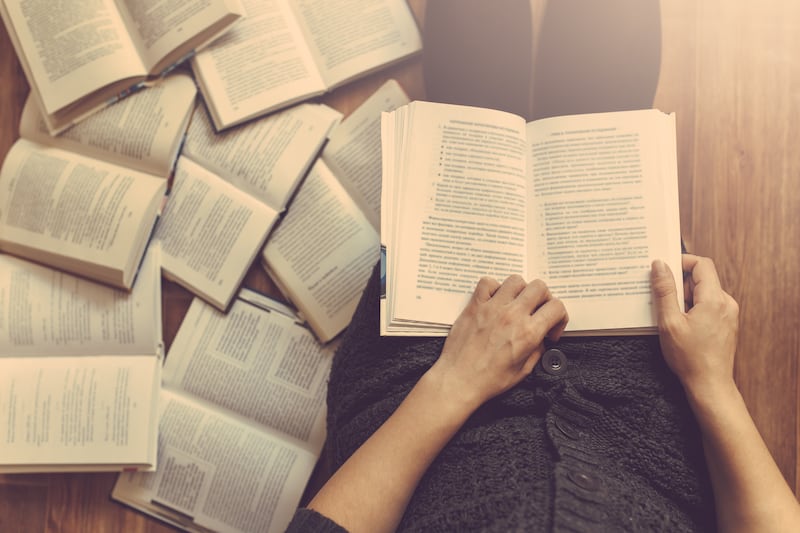 Una mujer lee un libro con varios libros en el suelo a su alrededor.
