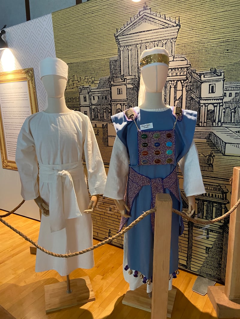 Un maniquí lleva una túnica blanca y otro maniquí lleva una túnica azul con 12 piedras de colores en la parte delantera, en el modelo del antiguo tabernáculo portátil de Moisés.