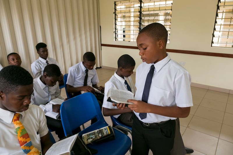 Un grupo de hombres jóvenes sentados en una clase del sacerdocio en Ghana. Un joven está de pie leyendo las escrituras.