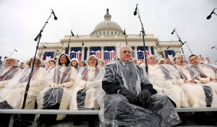 Director Mack Wilberg, in a dark suit, and choir members in beige coats with red plaid scarves wear clear rain ponchos as they sit on risers outside of the Capitol.
