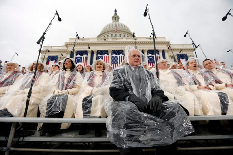 Director Mack Wilberg, in a dark suit, and choir members in beige coats with red plaid scarves wear clear rain ponchos as they sit on risers outside of the Capitol.