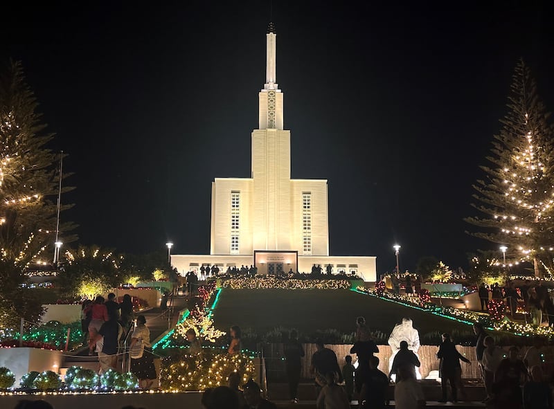 Los jardines del Templo de Hamilton, Nueva Zelanda, están iluminados con luces navideñas en esta foto de diciembre de 2025. En primer plano, los visitantes observan una escena del Nacimiento frente al templo.