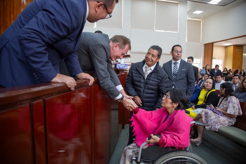 El élder Gary E. Stevenson, del Cuórum de los Doce Apóstoles, se inclina para estrechar la mano de una hermana en silla de ruedas y saludar a otros asistentes.