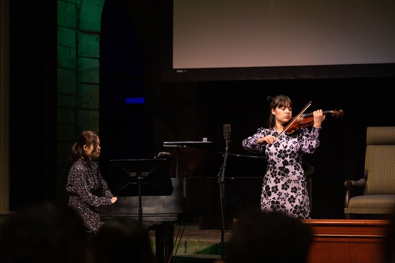 A young woman playing a violin next to another young woman playing piano during an Ensign College devotional.