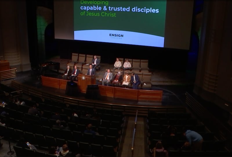 People in formal attire sitting in seats behind a pulpit for an Ensign College devotional.