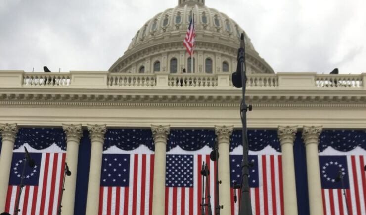 Choir members in beige coats with red plaid scarves wear clear rain ponchos as they sit on risers outside of the Capitol.