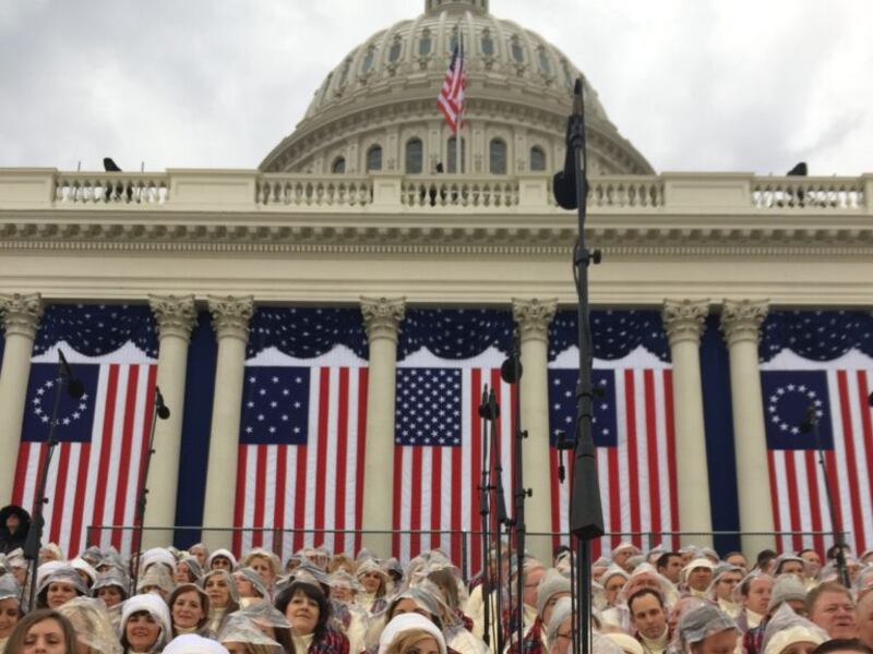 Choir members in beige coats with red plaid scarves wear clear rain ponchos as they sit on risers outside of the Capitol.