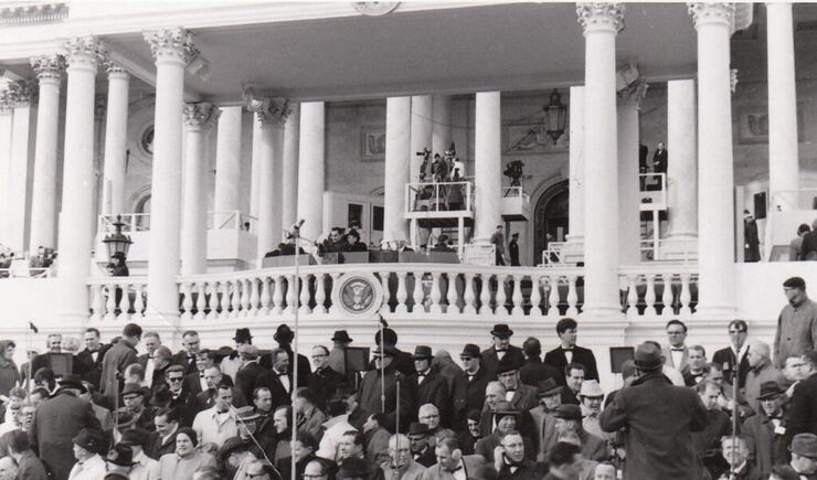 Tabernacle Choir members stand on risers as they prepare to sing outside a porch with tall white columns.