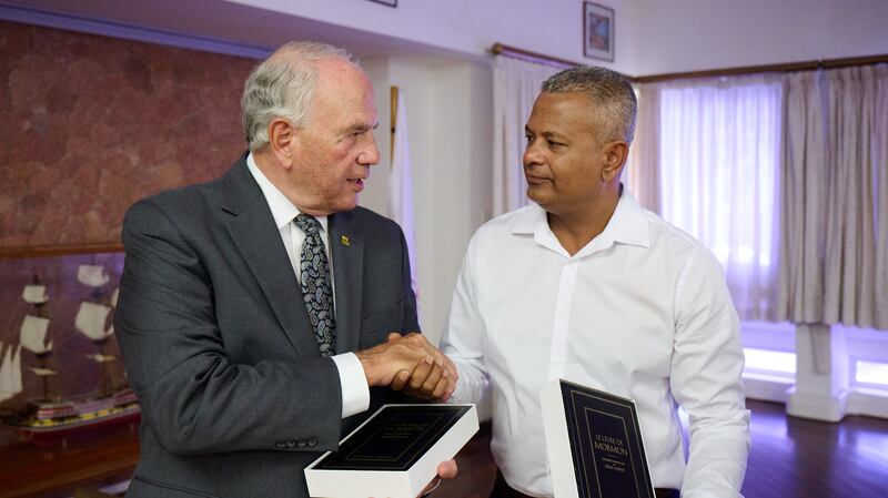 El élder Ronald A. Rasband estrecha la mano de Sebastien Pillay, el vicepresidente de Seychelles, mientras sostienen ejemplares del Libro de Mormón el 29 de octubre de 2025.